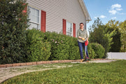 Photo of person using CRAFTSMAN string trimmer CMESTHOS912 along a garden walkway next to a house with red shutters.