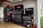 A garage scene featuring multiple CRAFTSMAN branded black tool chests and cabinets with silver handles, including a large tool chest, a rolling tool cart, and shelving units filled with various tools and supplies.
